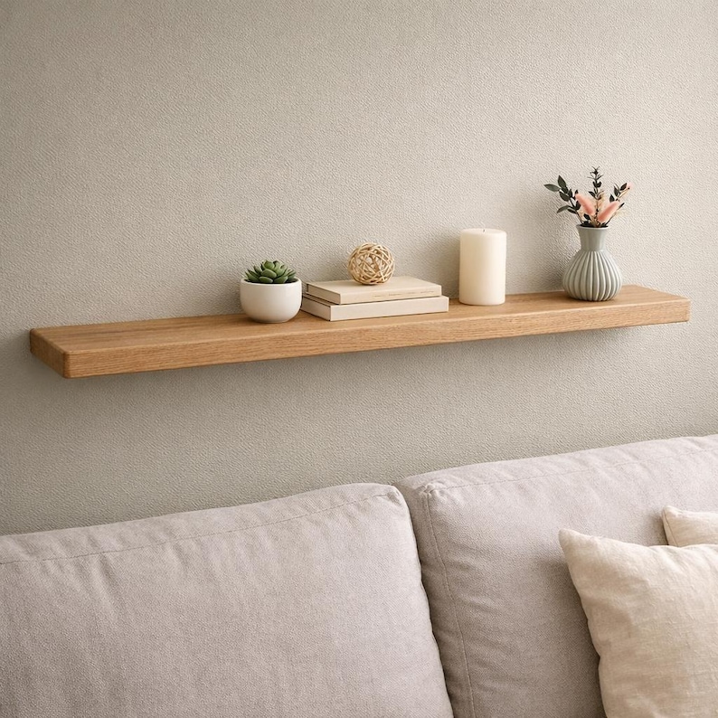 Puede incluir: Estante flotante de madera marr&oacute;n claro con varios objetos decorativos. El estante contiene una peque&ntilde;a suculenta en una maceta blanca, dos libros, una bola decorativa, una vela blanca y un peque&ntilde;o jarr&oacute;n con flores.