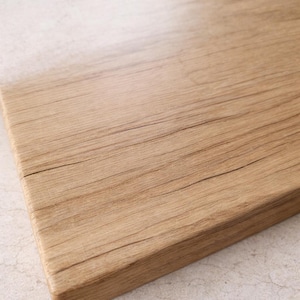 Puede incluir: Primer plano de una tabla de cortar rectangular de madera. La tabla es de color marr&oacute;n claro con vetas de madera visibles y grietas naturales. Los bordes son lisos y redondeados.