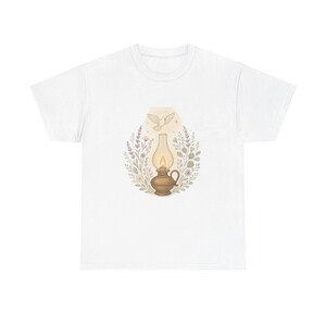 Op de afbeelding: Wit T-shirt met een aquarel illustratie. Het ontwerp toont een vintage olielamp met een vlam, een duif en bloemelementen in zachte kleuren. Het kunstwerk is gecentreerd op het T-shirt.
