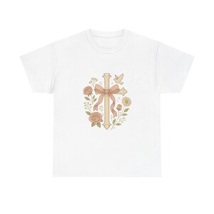 Może przedstawiać: Biały t-shirt z krótkim rękawem z centralną grafiką przedstawiającą beżowy krzyż ozdobiony delikatną różową kokardą, otoczony ilustracjami róż, drobnych kwiatów, zielonych liści oraz małego lecącego ptaka w stonowanych pastelowych barwach.