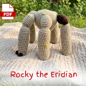 May include: A crocheted beige creature with six legs and a rounded body, resting on a textured blanket. The creature has a brown appendage and yellow accents. The text "Rocky the Eridian" is displayed in red.