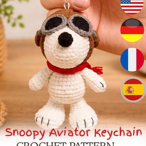 Könnte beinhalten: Gehäkelter Snoopy-Aviator-Schlüsselanhänger. Die Snoopy-Figur ist weiß und trägt eine braune Fliegermütze mit schwarzer Brille und einem roten Schal. Der Text "Snoopy Aviator Keychain CROCHET PATTERN" ist sichtbar.