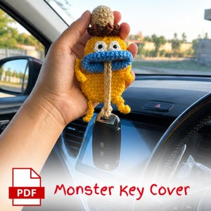 May include: A handmade yellow crochet monster key cover with a blue mouth, brown hat, and beige string. The key cover is holding a black car key. The text "Monster Key Cover" is visible in the image.