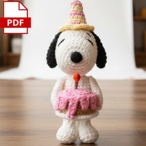 May include: A white crocheted dog figure with black ears and a black nose, wearing a yellow and pink striped party hat. The figure holds a small brown cake with pink frosting and a single orange candle on top, crafted in a soft yarn texture.