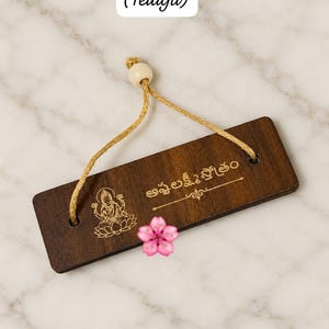Könnte beinhalten: Ein rechteckiges, dunkelbraunes Holzschild mit einer goldfarbenen Gravur einer Gottheit auf einem Lotus und dem Telugu-Text Ashtalakshmi stotram. Eine goldene Kordel mit einer cremefarbenen Perle ist zum Aufhängen angebracht.