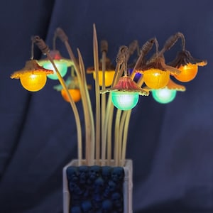May include: A decorative arrangement featuring multiple illuminated, mushroom-shaped lamps attached to thin, light-colored wooden stems. The lamps emit glowing orange, green, and blue light. The stems are held in a rectangular vase with a dark skull pattern.