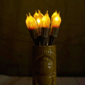 May include: A cluster of glowing LED tiki torches with flickering flame effects. The torches are arranged in a yellow ceramic tiki mug. The torches have a realistic flame appearance, creating a warm, inviting ambiance.