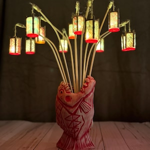 May include: A decorative arrangement featuring small, illuminated lanterns attached to bamboo sticks, displayed in a red fish-shaped vase. The lanterns have colorful patterns and red bases, creating a warm, inviting glow. The vase has a detailed fish design.