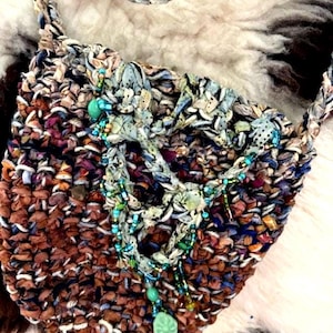 Puede incluir: Un bolso tejido a mano con una correa trenzada. El bolso presenta una mezcla de tiras de tela marrón, azul y morado. Adornado con cuentas turquesas y un colgante verde, descansa sobre una superficie texturizada blanca y marrón.