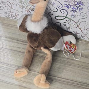 May include: A plush ostrich toy with a tan neck and legs, brown body, and white accents. The toy has a white collar and a red heart-shaped tag attached. The background features a patterned design with snowflakes.
