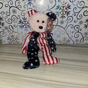 Könnte beinhalten: Ein Plüschteddybär mit rosa Gesicht, schwarzen Augen und Nase sowie einem rot-weiß gestreiften und blau-sternbesetzten Design. Der Bär hat ein Ty-Etikett an seinem Ohr befestigt. Der Bär ist ein Sammlerstück mit patriotischem Thema.