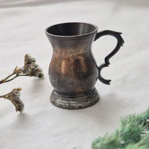 May include: A small, tarnished metal cup with a dark, weathered patina and a curved, ornate handle. The vessel features a flared rim and a sturdy, circular base, showing signs of age and oxidation across its metallic surface.