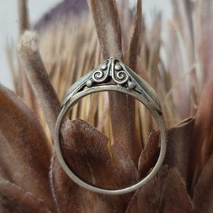 Op de afbeelding: Een zilveren ring met een puntig, verhoogd middendeel versierd met ingewikkeld krulwerk en kleine bolvormige accenten. Het metaal heeft een licht geoxideerde afwerking, waardoor de gedetailleerde gravures op het bovenste deel van de ring opvallen.