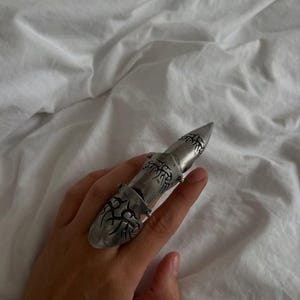 May include: A silver-toned articulated finger ring featuring a segmented design that covers the length of the finger. The metal surface is engraved with dark, intricate tribal-style patterns on each of the three connected sections.