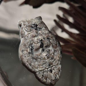 May include: A carved stone owl figurine featuring a mottled gray, brown, and black speckled pattern. The owl is depicted with detailed feathers, prominent eyes, and a textured surface, showcasing natural stone variations throughout the entire piece.