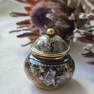 Può includere: Un piccolo vasetto rotondo in ceramica nera con coperchio, decorato con illustrazioni colorate e dettagliate di figure classiche in un giardino. Il vaso presenta un bordo dorato, una base dorata e un coperchio a cupola con pomello dorato.