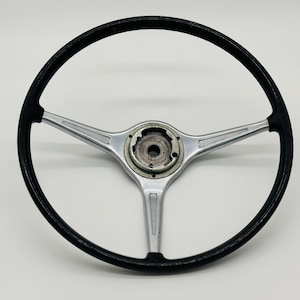 May include: A classic steering wheel featuring a black rim and a three-spoke design. The silver spokes have a textured pattern, and the central hub is metallic. This vintage car part is designed for a vehicle.