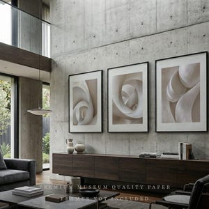 Puede incluir: Tres impresiones artísticas abstractas enmarcadas con tonos neutros cuelgan de una pared de hormigón. La obra de arte presenta formas orgánicas arremolinadas en tonos beige y blanco. Un mueble y un sofá de madera oscura están en primer plano.