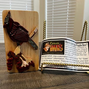 May include: A wooden puzzle box featuring a carved cardinal bird design. The bird is primarily red with accents of white and iridescent blue-green. The box includes a label with instructions and the text "Carver Dan".