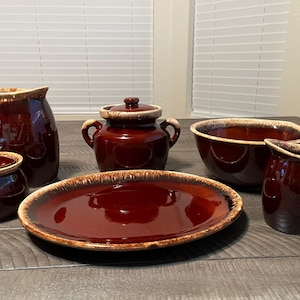May include: A collection of vintage brown ceramic kitchenware, including a pitcher, a covered sugar bowl, a casserole dish, a serving bowl, a platter, and a small creamer. The pieces have a glossy finish and a two-tone design.