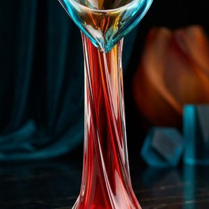 May include: A tall glass vase featuring a twisted, fluted body that transitions from a deep red base to a vibrant turquoise and amber rim. The opening is wide and irregularly shaped, with clear glass edges that highlight the swirling color pattern.