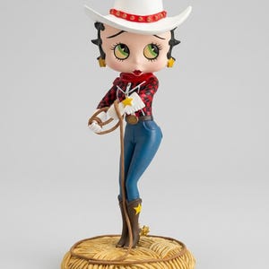 May include: A figurine of a cartoon character in a cowgirl outfit. The figure wears a white cowboy hat with a red band, a red and black plaid shirt, blue jeans, and brown boots. The figurine stands on a hay bale base.