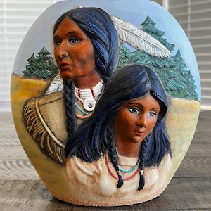 May include: A ceramic vase with a relief of two Native American figures. The figures have dark hair and are adorned with necklaces. The background features a blue sky, green trees, and a large white feather. The vase has a rounded shape.