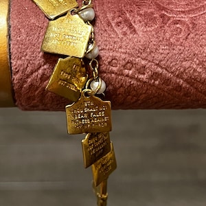 May include: A gold-colored charm bracelet featuring small rectangular charms with engraved text and faux pearls. The charms are connected by a delicate chain. The bracelet is displayed against a pink velvet background.