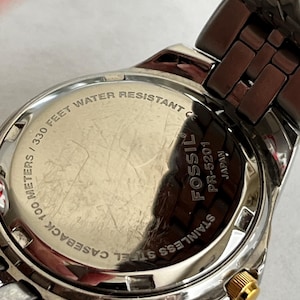 May include: Close-up of a stainless steel Fossil watch. The watch back is visible, displaying text that reads "330 FEET WATER RESISTANT" and "STAINLESS STEEL CASEBACK 100 METERS". The watch has a silver and gold-tone metal band.