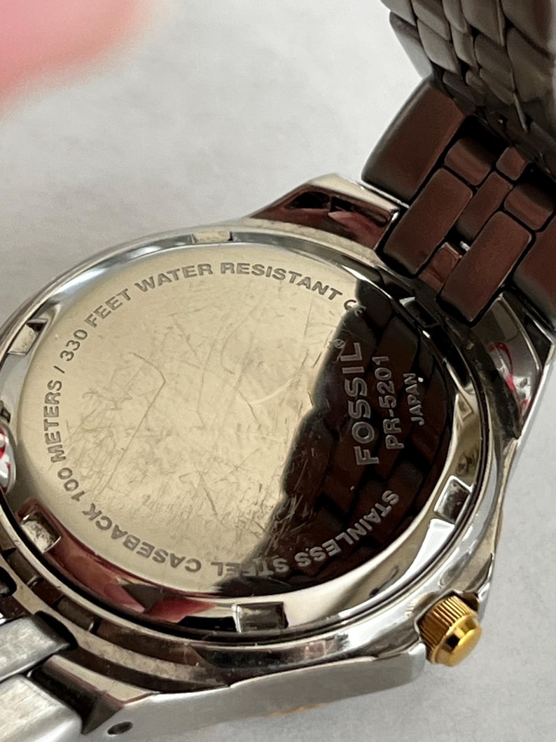 May include: Close-up of a stainless steel Fossil watch. The watch back is visible, displaying text that reads "330 FEET WATER RESISTANT" and "STAINLESS STEEL CASEBACK 100 METERS". The watch has a silver and gold-tone metal band.