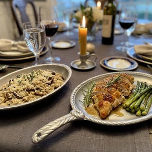 May include: A beautifully set dining table with a meal of sliced chicken, asparagus, and risotto. The food is served on decorative oval platters. Wine glasses, candles, and plates complete the elegant setting.
