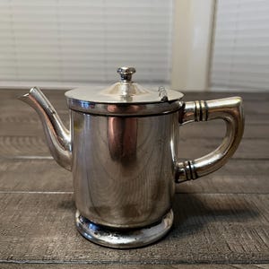 May include: A small, silver-colored teapot with a lid and handle. The teapot has a spout for pouring and sits on a round base. The handle has a curved design. The teapot is made of metal and has a shiny finish.