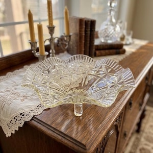 May include: Clear glass serving dish with a starburst and diamond pattern. The dish has a scalloped edge and sits on a short pedestal. The dish is on a wooden surface, with candles and books in the background.