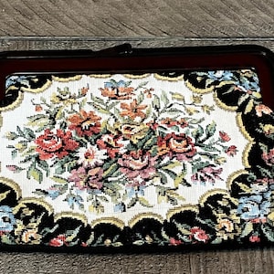 May include: A vintage tapestry clutch with a black frame and clasp. The central design features a floral arrangement in shades of red, orange, pink, and blue on a white background. The edges are adorned with black and gold floral patterns.