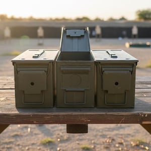 May include: Three olive drab metal ammo cans are displayed on a wooden table. The center can is open, revealing its interior. The background shows a shooting range with targets. The cans have hinged lids and secure latches.
