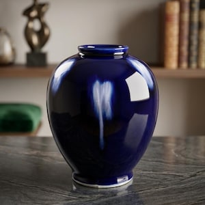 May include: A glossy, deep blue ceramic vase with a rounded body and a narrow neck. The vase has a white base and a reflective surface, with light reflecting off the curves. The vase is a decorative home decor item.