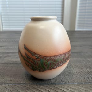 May include: A white ceramic mug with a black handle and the text "But First, Coffee" in a bold, black font. The mug is set against a blurred background, suggesting a cozy setting.