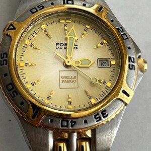 May include: A Fossil watch with a gold and silver-tone metal band and a gold face. The watch face displays the Wells Fargo logo and the text "Fossil 100 M RS". The watch has a date display at the 3 o'clock position.