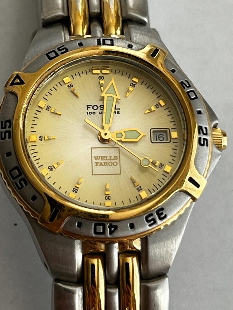 May include: A Fossil watch with a gold and silver-tone metal band and a gold face. The watch face displays the Wells Fargo logo and the text "Fossil 100 M RS". The watch has a date display at the 3 o'clock position.