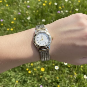 May include: A silver and gold-toned wristwatch with a white face and black numerals. The watch has a gold crown and a silver, mesh-style band. The watch is on a wrist, with a green and yellow floral background.