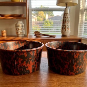 May include: Two oval-shaped, vintage bowls with a speckled red and black pattern. The bowls are made of a durable material and sit on a wooden table. The bowls are part of a kitchenware set.