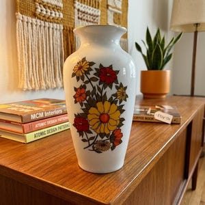 May include: A white ceramic vase with a floral design featuring yellow, orange, and red flowers. The vase has a gold rim and is displayed on a wooden surface. The vase is a decorative item.