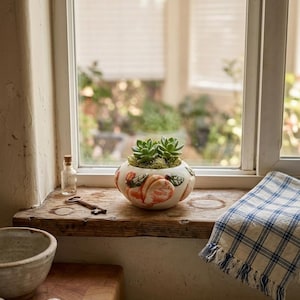 May include: A small, round ceramic planter with a peach design, holding green succulents, sits on a wooden windowsill. A small glass bottle, a key, and a plaid blue and white cloth are also on the sill. A rustic kitchen scene.