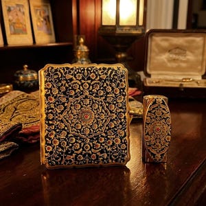 May include: A rectangular compact case and a matching cylindrical lipstick holder, both featuring a dark blue background with intricate gold, red, and white floral patterns. The items have scalloped edges and a metallic finish, displaying detailed craftsmanship.