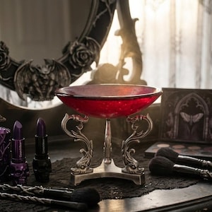Puede incluir: Un cuenco de cristal rojo sobre un soporte plateado ornamentado. El cuenco está sobre una superficie oscura con brochas de maquillaje, pintalabios y un espejo en el fondo. El estilo es gótico y vintage.