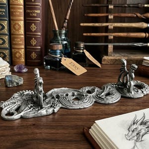 May include: A silver-toned metal dragon-shaped pen rest featuring two upright dragon figures with wings and coiled tails. The detailed sculpture has a textured, metallic finish and is designed to hold writing instruments horizontally between the two figures.