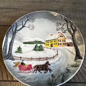 May include: A decorative ceramic plate featuring a winter scene. The plate depicts a yellow house with a red roof, a horse-drawn sleigh, and snow-covered trees. The plate has a raised relief design and is bordered by a silver rim.
