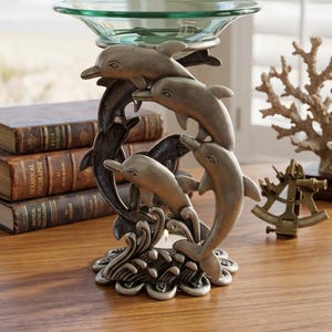 May include: A silver-toned metal candle holder featuring a sculptural base of three leaping dolphins emerging from stylized waves. The structure supports a clear, circular glass bowl at the top, designed to hold a small candle or tealight.