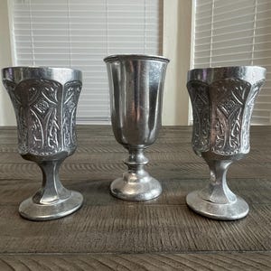 May include: Three silver-colored goblets with ornate designs and pedestal bases. The two outer goblets have a detailed, embossed pattern, while the central one is smooth. They are displayed on a wooden surface, suggesting a vintage or antique theme.