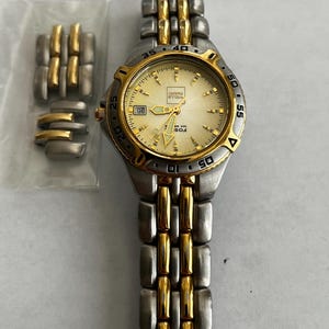 May include: A silver and gold-toned wristwatch with a cream-colored face. The watch has a date display at the 9 o'clock position and a gold bezel. Additional watch links are in a plastic bag.